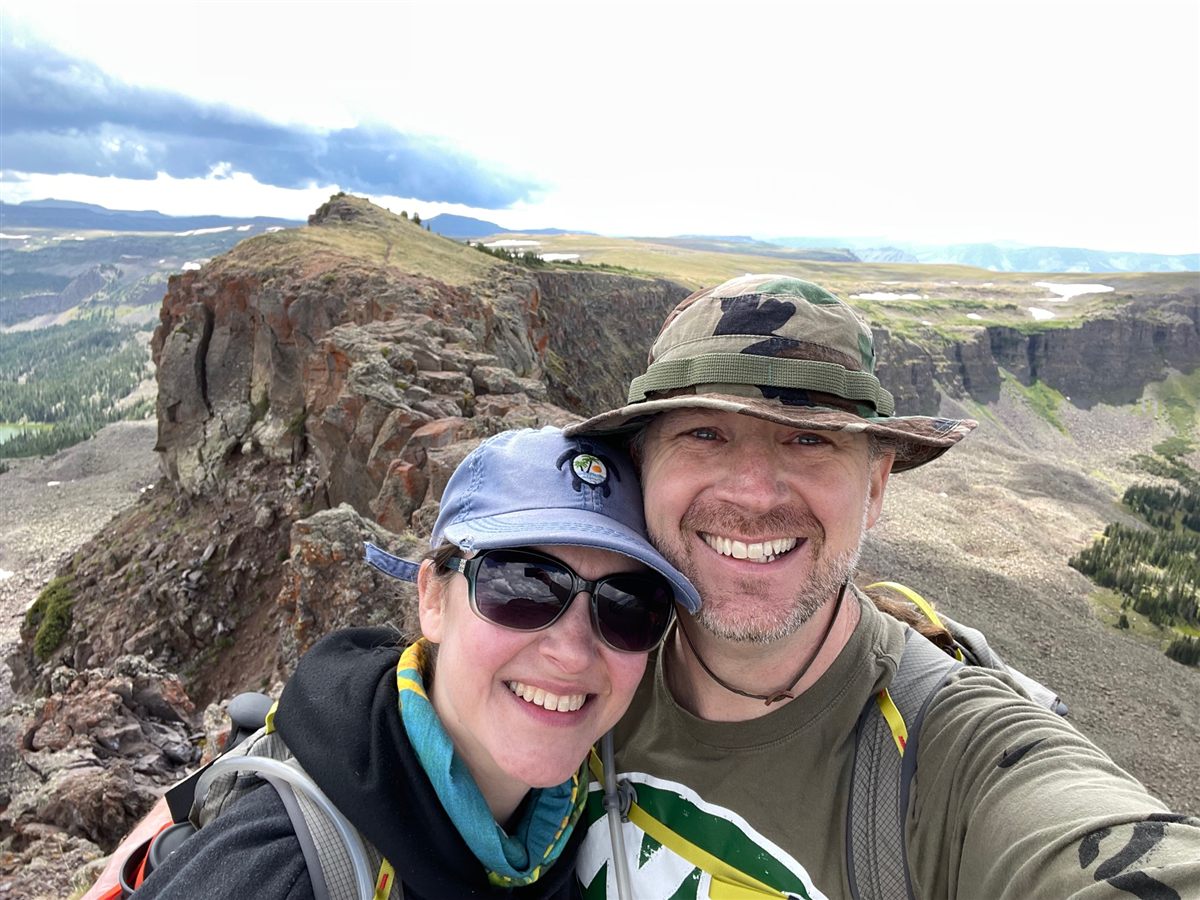 Alvin and his wife smiling on a rocky mountain ridge high above the valley